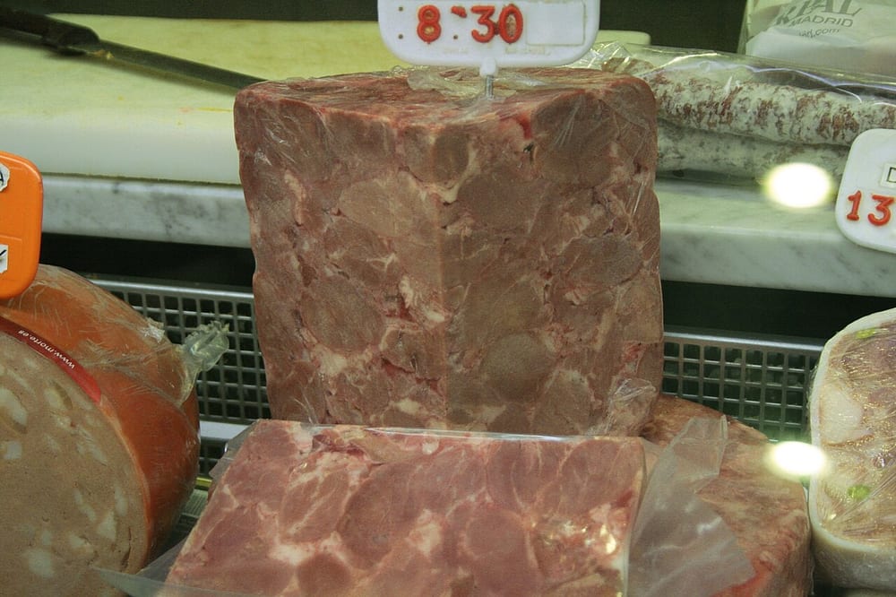 A cut terrine of head cheese: irregular chunks of dark and pale meat suspended in clear amber jelly. Nothing here is ground and nothing is emulsified — the set is gelatin cooked out of the animal's own connective tissue