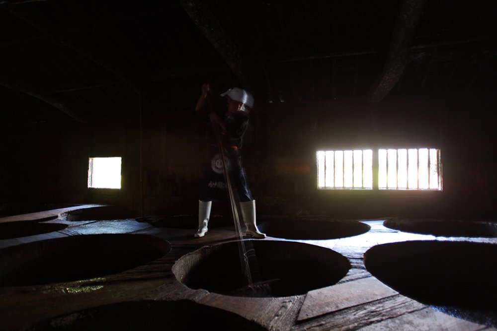 A soy sauce brewery interior, lit only by high windows, with a worker stirring the moromi mash with a long wooden paddle