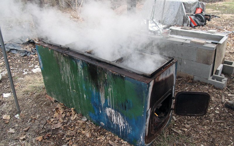 A homemade maple evaporator built from an upright filing cabinet, boiling hard with steam rolling off a shallow pan on top; the firebox door is swung open at the bottom showing burning wood inside