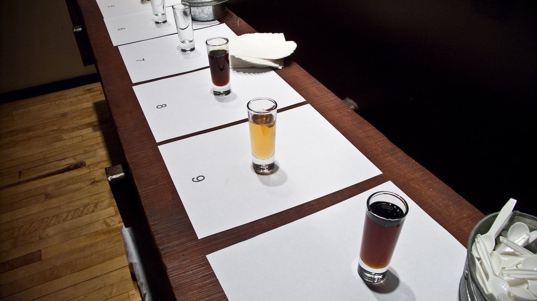 A maple tasting flight — numbered tasting glasses set out on a table, the syrup in them running from near-black through deep amber to pale gold, with spoons in a tin alongside