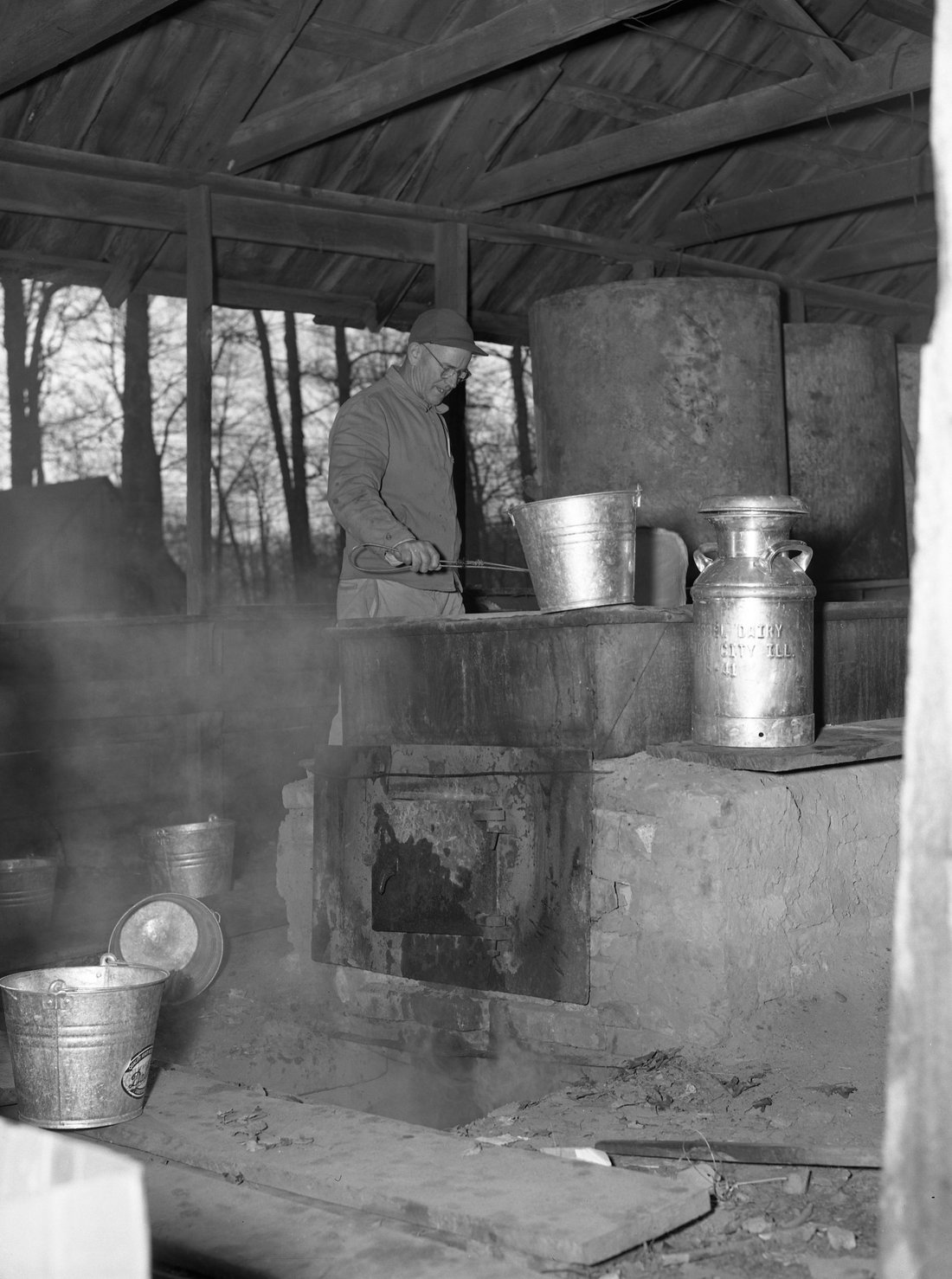 A sugarmaker tending a wood-fired arch in an open-sided shack, steam drifting off the pan, gathering buckets and a milk can standing by — the long reduction where the flavor is actually created