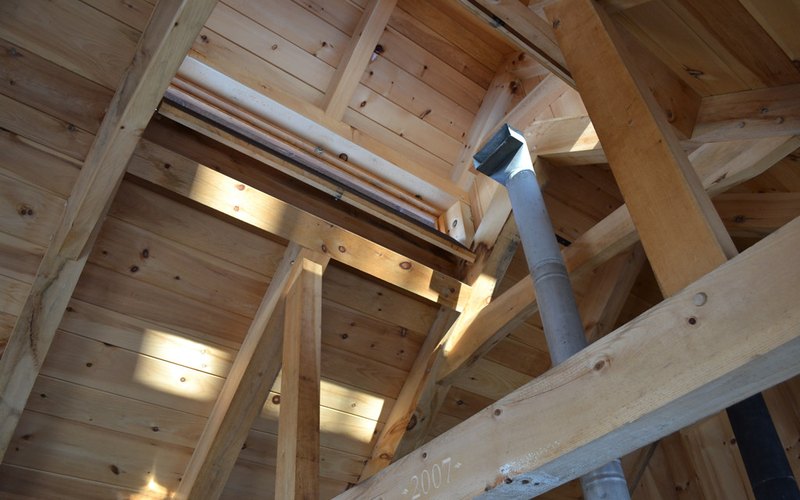 The inside of a timber-framed sugarhouse roof looking up: heavy rafters and collar ties converging on a long rectangular steam vent open to daylight along the ridge