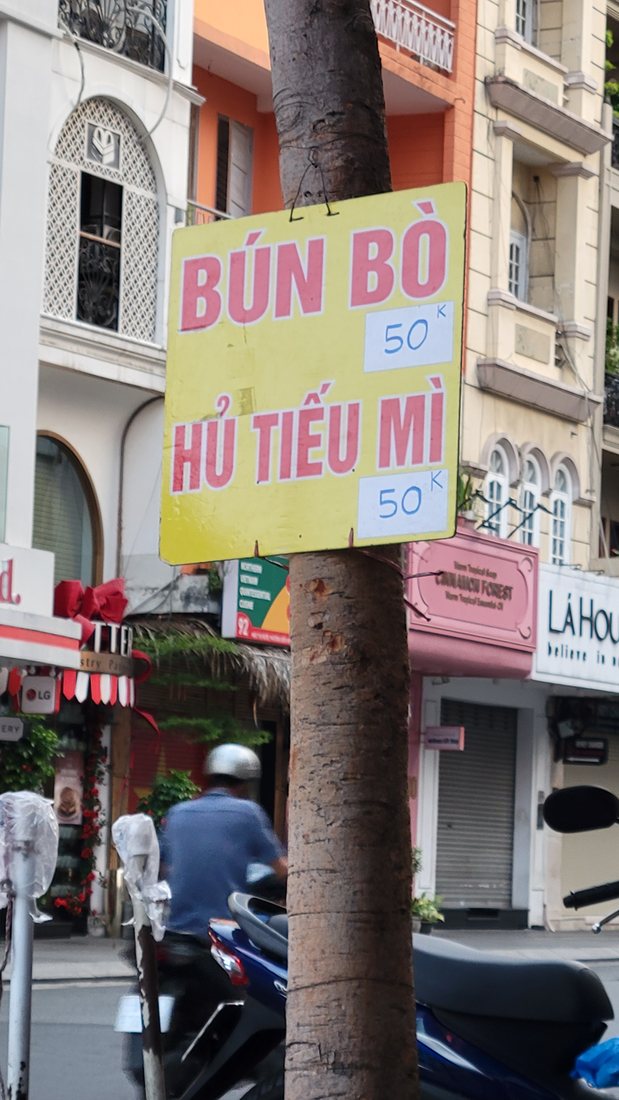 The street I ate it on — a BÚN BÒ / HỦ TIẾU MÌ board, prices in the fifties, a pole and a wall and no shopfront to speak of
