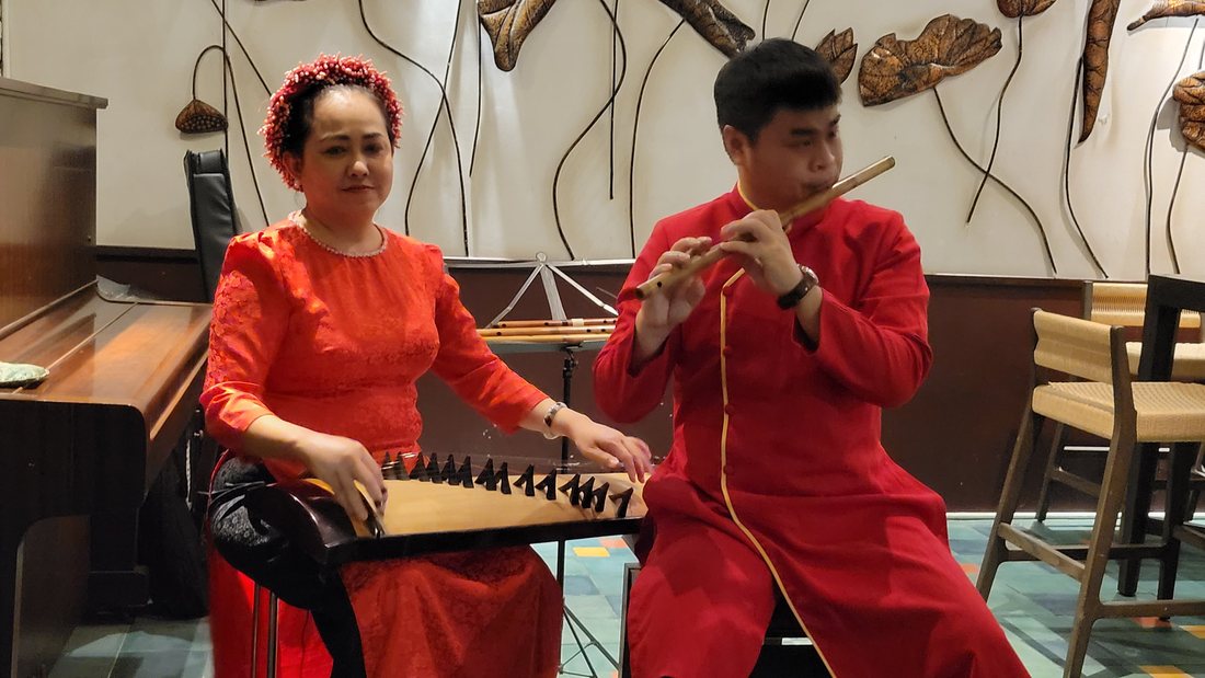 Two musicians in red áo dài at the side of the room — a đàn tranh on her lap, a flute in his hands