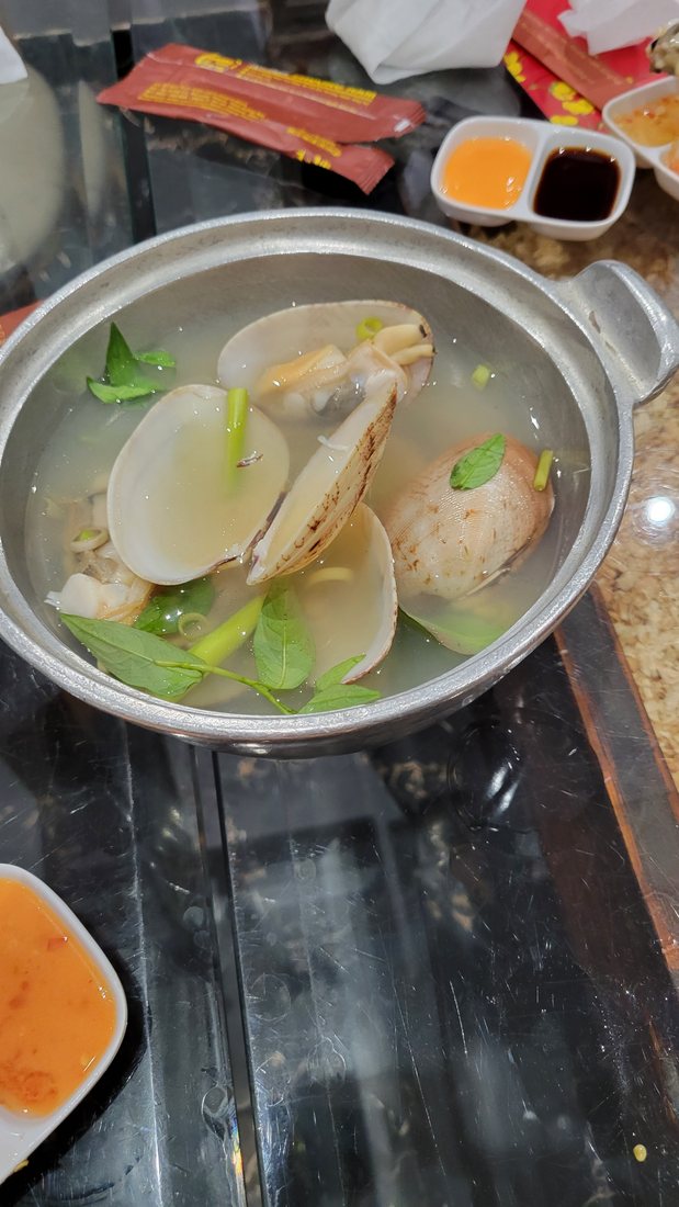Large clams, opened, in a clear broth with herbs and scallion, in an aluminium pot over a burner — with a two-part chilli-salt and dark-sauce dip alongside