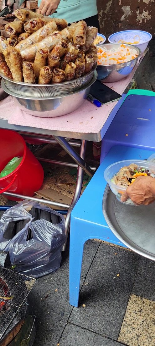 The other end of the operation, four seconds later: a colander mounded with fried chả giò, a steel bowl of đồ chua, a bowl being built on a plastic stool — and, under the table, the bags of charcoal