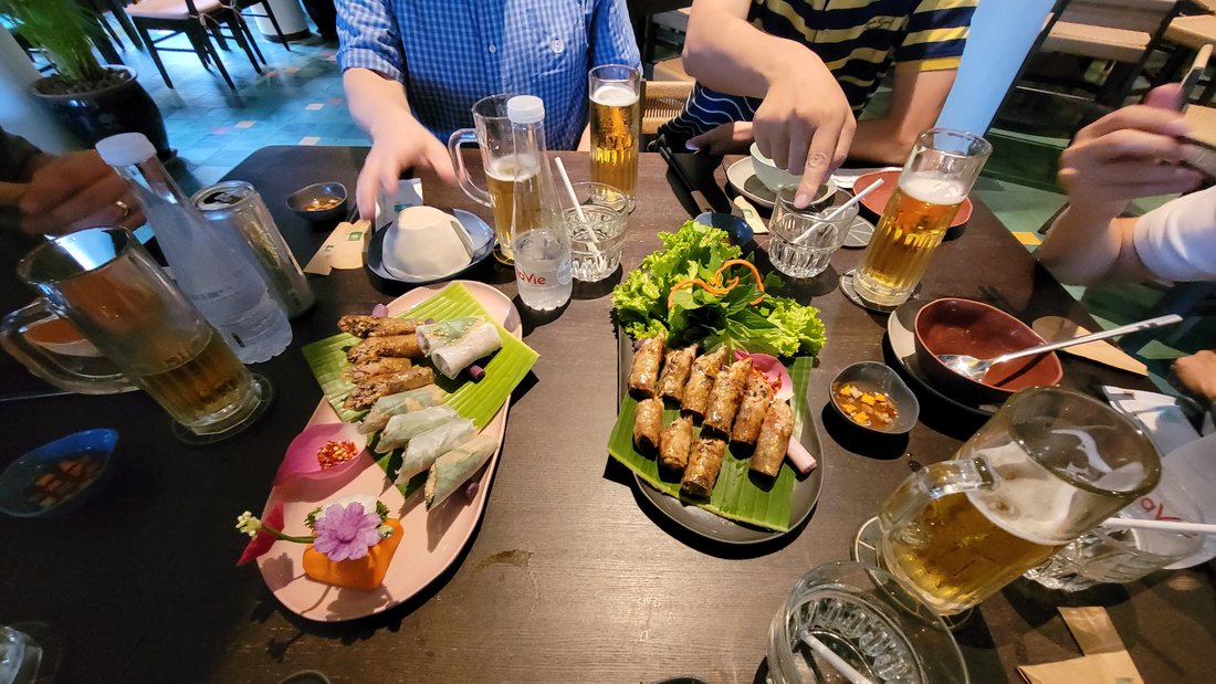 A long banana-leaf platter of grilled skewers with fresh herbs and lettuce laid down the middle of it, dipping bowls at either end, and a glass of beer per person