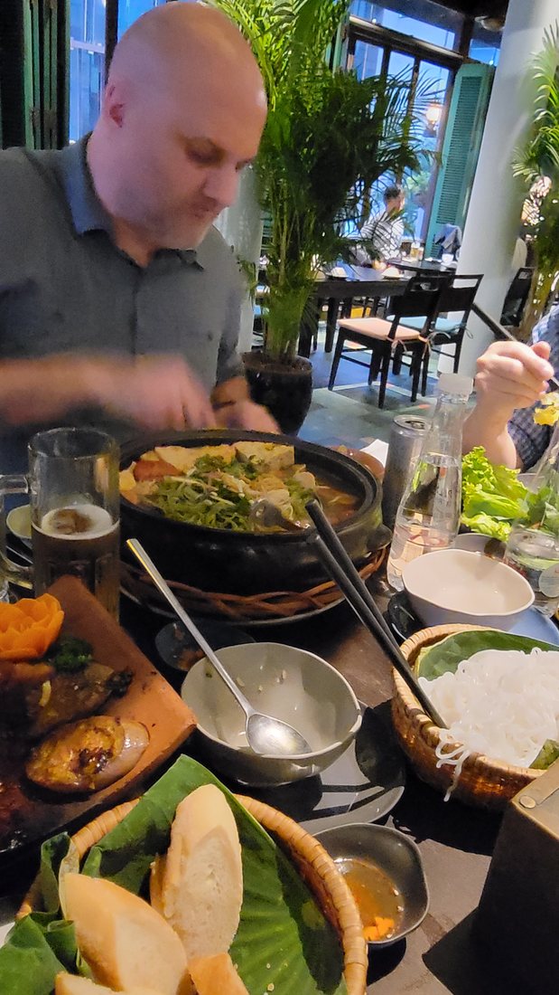 A black clay pot of noodles and greens over its own burner, with a basket of split bánh mì beside it