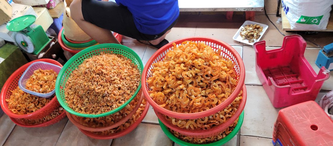 Three graded baskets of dried shrimp — the curl, the segmented shell and the tail are all clearly visible, sorted small to large, with the vendor's balance scale at the left of frame