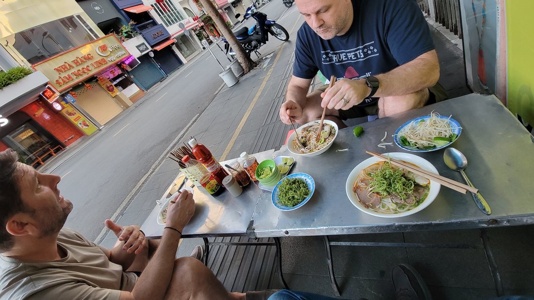 Phở at a steel pavement table, ten past six in the morning. Low stools, a shared table, and the bowl in front of you before you have finished sitting down