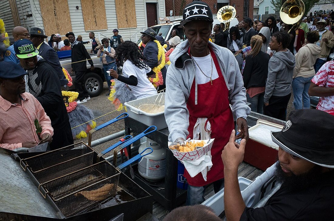 A vendor in a red apron handing food to a customer from a curbside cooker during a second line parade